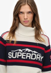 Młoda kobieta z długimi brązowymi włosami, ubrana w biało-granatowo-czerwony sweter z golfem w paski z napisem "SUPERDRY" oraz wzorem gór.