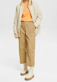 Beige bomberjacka över en orange T-shirt, tillsammans med khakifärgade, korta cargo-byxor och krämfärgade ankelstövlar med tjock sula.