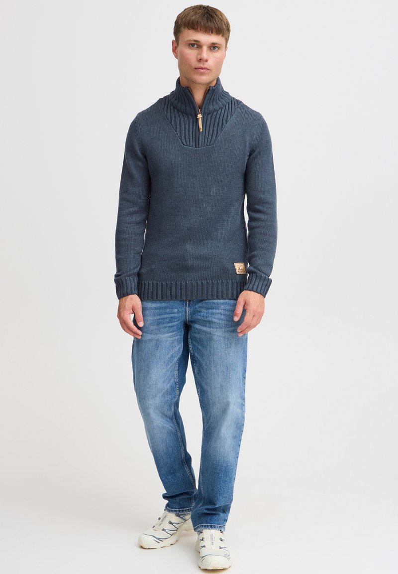 Marineblauer gestrickter Pullover mit hohem Kragen, Rippstruktur, ausgestattet mit einem Viertelreißverschluss und Logo-Patch. Kombiniert mit hellblauen Jeans und Sneakers.