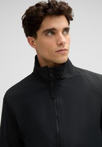 Schwarze, leichte Jacke mit hohem Kragen, ausgestattet mit einem Reißverschluss vorne und strukturiertem Stoff. Markenlogo sichtbar am Reißverschluss-Zipper.