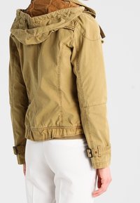 Kaki jacka med huva, elastisk midja och knappade ärmslut. Har en texturerad tyg och synligt sömnadsdetaljer.