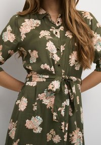 Femme portant une robe floral olive verte avec des fleurs roses et bleu clair, des boutons noirs et une ceinture à nouer, avec de longs cheveux ondulés visibles.