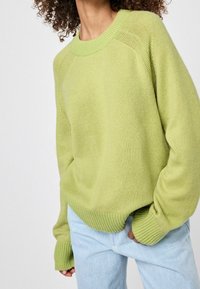 Pull en maille vert clair avec un col arrondi, des poignets et un bas côtelés, et une coupe décontractée. Associé à un jean bleu clair.