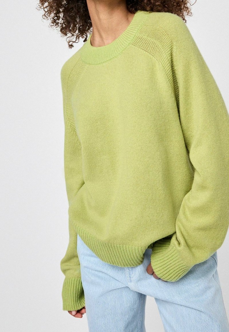 Pull en maille vert clair avec un col arrondi, des poignets et un bas côtelés, et une coupe décontractée. Associé à un jean bleu clair.