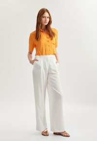 Oranje overhemd met korte mouwen en een textuur, gecombineerd met hoge witte wijde broekspijpen en metallic bandjesandalen.