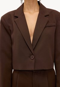 Personne portant un blazer court marron foncé avec un bouton fermé et un pantalon taille haute assorti, laissant apparaître le décolleté et les clavicules.