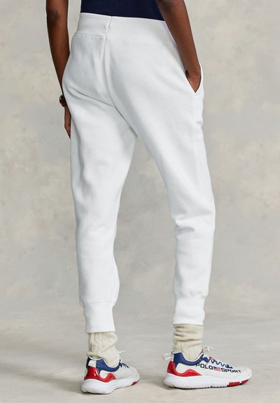 Άτομο που φοράει λευκό παντελόνι φόρμας, κρέμ καλτσοδέτες από πλεκτό καλώδιο και λευκά, κόκκινα και ναυτικά αθλητικά παπούτσια Polo Sport, όρθιο με τα χέρια στις τσέπες.