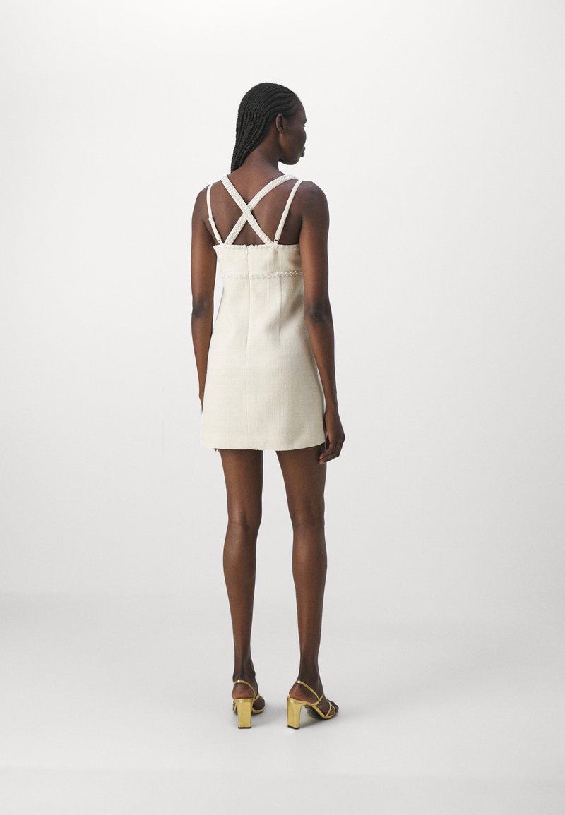 Beige Kleid in Knielänge mit überkreuzten weißen Trägern, strukturiertem Stoff und taillierter Silhouette. Kombiniert mit gelben Absatzsandalen.