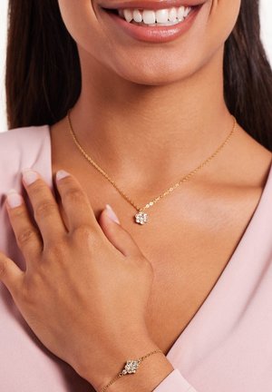 Gouden ketting met een florale kristalhanger, gecombineerd met een bijpassende armband met vergelijkbaar kristalontwerp. Huidtint zichtbaar.