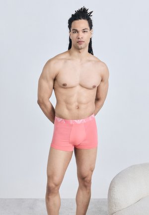 Homme musclé aux dreadlocks, torse nu, portant un boxer corail avec une ceinture à logo, debout devant un fond gris clair.