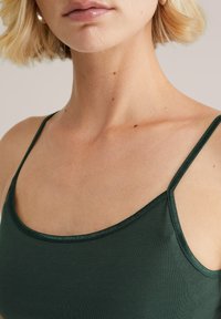 Blonde sieviete ar īsiem matiem, tērpusies tumši zaļā spageti stila topā, atklājot kaklu, plecus un augšējo krūtis.