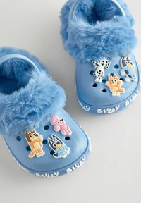 Blaue Clogs mit flauschiger blauer Innenausstattung, ausgestattet mit Cartoon-Charms in verschiedenen Farben und Belüftungslöchern auf der Oberfläche.
