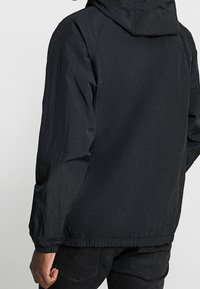 Man med svart huva jacka och svarta jeans, sedd bakifrån mot en enfärgad ljus bakgrund.