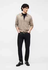 Άνδρας με σγουρά μαλλιά, φορώντας μπεζ ζακέτα με κουμπιά πάνω από μαύρο πουκάμισο, μαύρο τζιν και μαύρα παπούτσια, όρθιος με τα χέρια στις τσέπες.