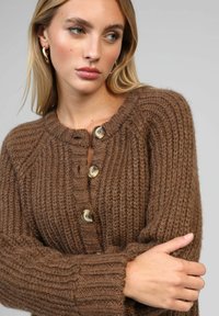 Gilet en tricot marron doté d'un col rond, d'une texture côtelée et de quatre grands boutons à l'avant. Tissu doux et épais.