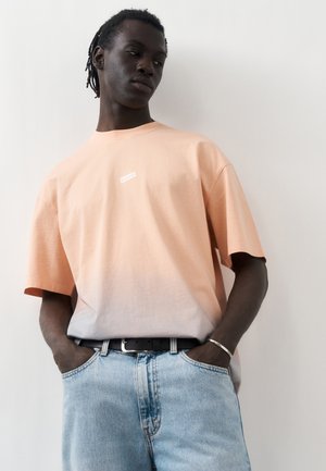 Mlad moški s temnejšo poltjo in dredlji nosi majico z gradientom iz breskve v belo, svetlo modre kavbojke, črni pas in srebrno zapestnico, roke v žepih.