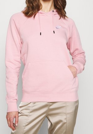Femme portant un sweat à capuche Nike rose clair avec cordons et un pantalon en satin beige, main dans la poche avant, sur un fond uni.