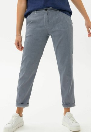 Person trägt hochgekrempelte, eng anliegende graue Hose mit Muster, vorne mit Knopf und Reißverschluss, kombiniert mit weißen Plateausneakern.