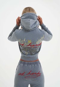 Grijze hoodie met een capuchon, voorzien van kleurrijke tekst en grafische ontwerpen op de achterkant. Gecombineerd met grijze joggers, die ook rode borduursels tonen.