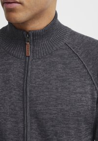 Temno siva zip-up jopica z ribanim ovratnikom in subtilno teksturo. Ima eleganten zadrgo z majhno usnjeno oznako z napisom "BLEND."
