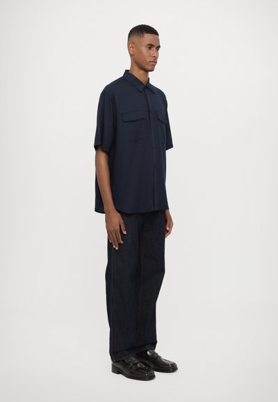 Marineblå kortermet skjorte med doble brystlommer, kombinert med mørk denimbukser og svarte loafers. Enkel design, myk tekstur.