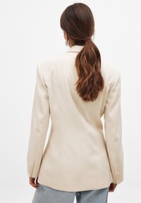 Beiger taillierter Blazer mit strukturierten Schultern, langen Ärmeln und drei-Knopf-Details an den Manschetten. Glattes Material mit einer figurschmeichelnden Silhouette.