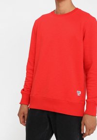 Persona vestida con un sudadera roja brillante con puños y dobladillo acanalados, combinada con pantalones negros, etiqueta pequeña blanca que dice "WHY RED" visible.