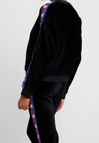 Veste en velours côtelé noire avec des poignets côtelés, présentant des rayures latérales rouges et bleues avec des accents de logo sur les manches.