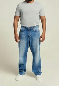 Camisa de algodón gris claro combinada con unos vaqueros de corte relajado en un tono azul desgastado. El conjunto incluye unas zapatillas deportivas blancas y cuenta con un diseño clásico de cinco bolsillos.