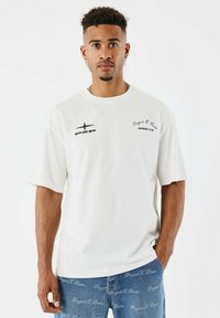 Joven con cabello corto y rizado que lleva puesta una camiseta blanca con logos negros y jeans azules bordados con el texto "Project X Paris".