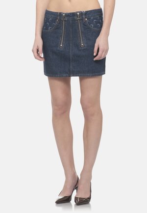 Donna che indossa una minigonna in denim scuro con cerniere verticali e tasche anteriori a fantasia, abbinata a décolleté scure e appuntite.