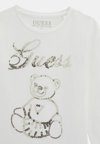 Camicia di cotone bianca con testo "Guess" in paillettes argento e un orsetto sorridente che tiene un pallone da basket, cucito sul davanti.