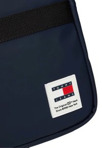 Granatowa torba o teksturowanej powierzchni, z czarnym paskiem i białą łatą z logo "TOMMY JEANS" w kolorze czerwonym i niebieskim.