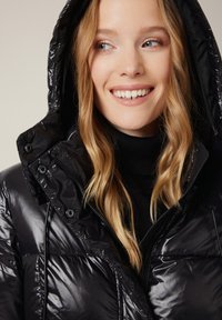 Uśmiechnięta kobieta z luźnymi blond włosami, w błyszczącej czarnej pikowanej kurtce z kapturem i czarnym swetrze z golfem.
