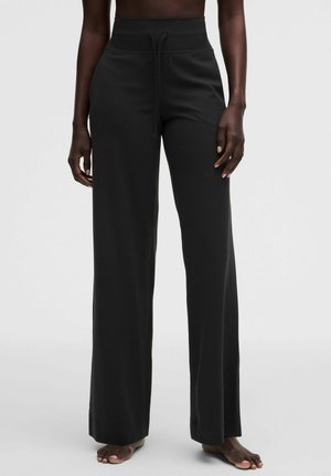 Personne portant un pantalon noir taille haute à jambes larges avec un cordon à l'avant, debout pieds nus sur un fond clair uni.