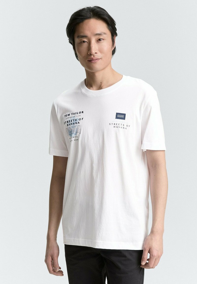 Hvid bomulds t-shirt med navy grafisk tekst på fronten, herunder brandnavn og placering, med rund hals og korte ærmer.