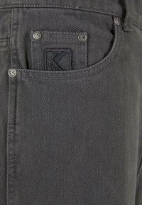 Grå denimjeans har en baklomme med en svart brodert logo og metallknapper, som viser et teksturert stoffmønster.