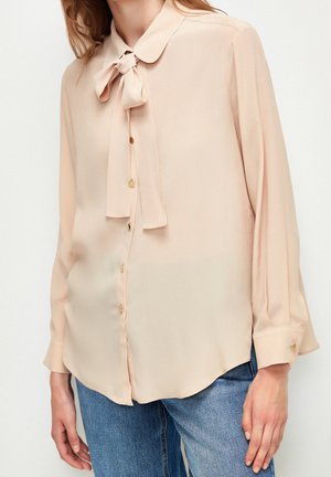 Femme portant une blouse beige clair à manches longues avec un nœud au col et des boutons dorés, associée à un jean en denim bleu.