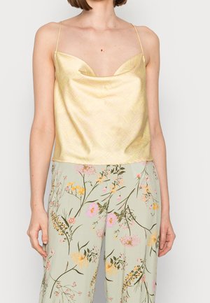 Frau trägt ein hellgelbes Satinkleid mit dünnen Trägern und weit geschnittene, hellgrün mit Blumenmuster bedruckte Hosen.