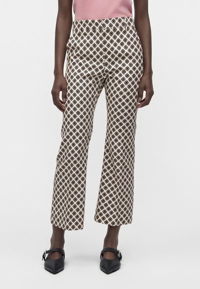 Femme portant un pantalon large crème avec un motif géométrique noir, un haut rose et des chaussures noires à bout pointu avec des boucles, debout sur fond blanc.
