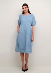 Gai zila džinsu kleita ar kvadrātveida izgriezumu, pūkainām piedurknēm, sānu kabatām un daudzslāņu apakšmalu. Valkāta ar melnām augstpapēžu kurpēm.