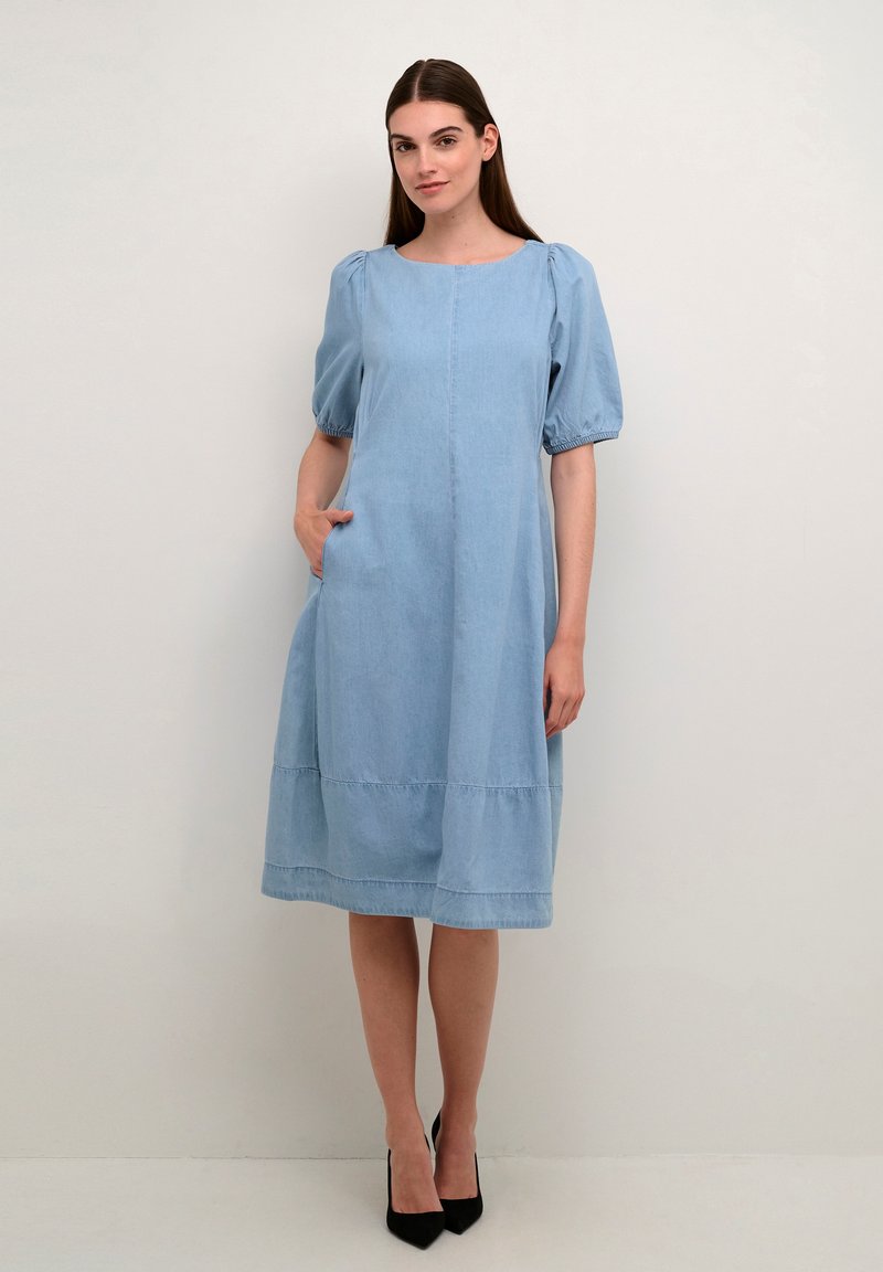 Gai zila džinsu kleita ar kvadrātveida izgriezumu, pūkainām piedurknēm, sānu kabatām un daudzslāņu apakšmalu. Valkāta ar melnām augstpapēžu kurpēm.