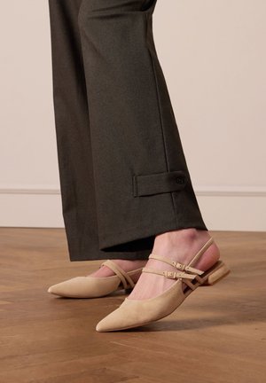 Beige suede slingback shoes with pointed toes, featuring double ankle straps and a low block heel, paired with dark tailored trousers.