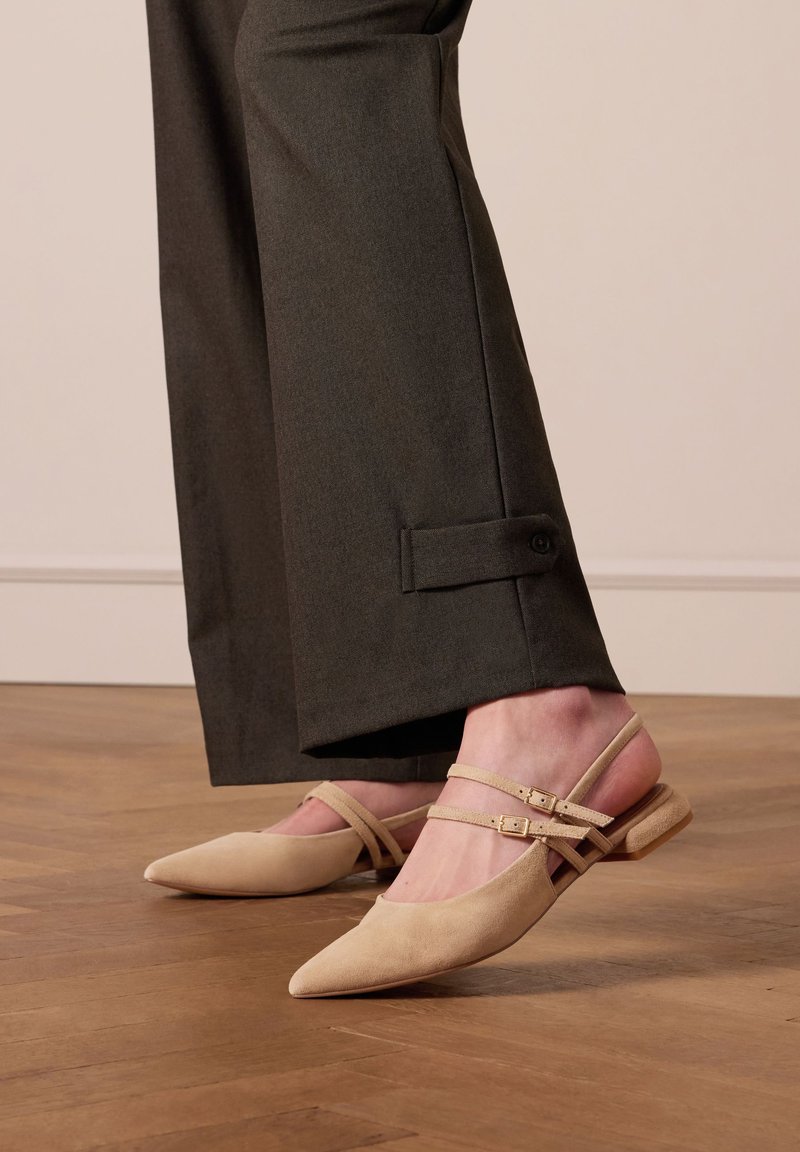 Beige suède slingback schoenen met puntige neuzen, voorzien van dubbele enkelbandjes en een lage blokhak, gecombineerd met donkere, op maat gemaakte broek.