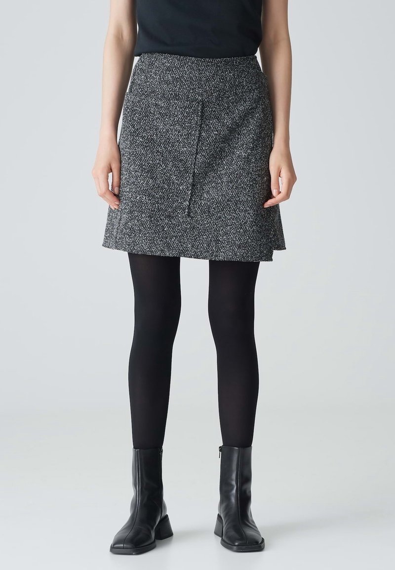 Femme portant une jupe en tweed grise, des collants noirs et des bottines noires, debout, les mains détendues le long du corps, devant un fond uni.
