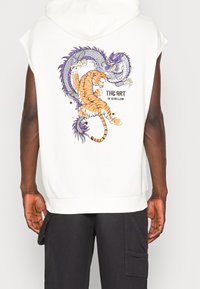 Vit ärmlös hoodie med en livfull grafisk bild av en tiger och en drake med blomsteraccentuerade detaljer, texten "THE ART OF REBELLION."