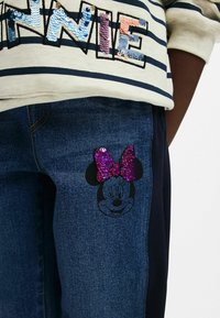 Plave traperice s crnim obrisom Minnie Mouse i sjajnom ljubičastom šljokičastom mašnom. Prugasta svijetla majica ukrašena šarenim šljokicama.