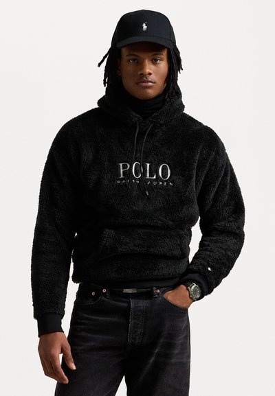 Musta fleece-huppari, jossa on "POLO Ralph Lauren" -logo, varustettu kiristettävällä hupulla, käytetty mustan poolopaidan päällä, yhdistettynä mustiin farkkuihin ja kelloon.