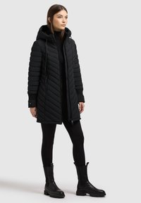 Kobieta ubrana w czarny pikowany płaszcz z kapturem, czarne legginsy i czarne botki, stojąca na tle jednolitego jasnoszarego tła.