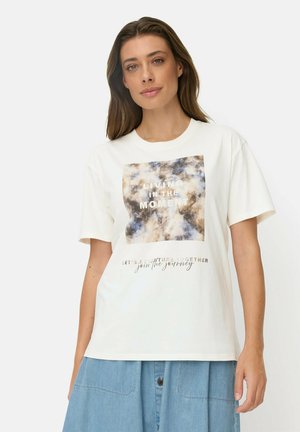 Vrouw met lang bruin haar die een wit T-shirt draagt met een blauw en bruin afbeelding en tekst, en een lichtblauwe spijkerrok met knopen.